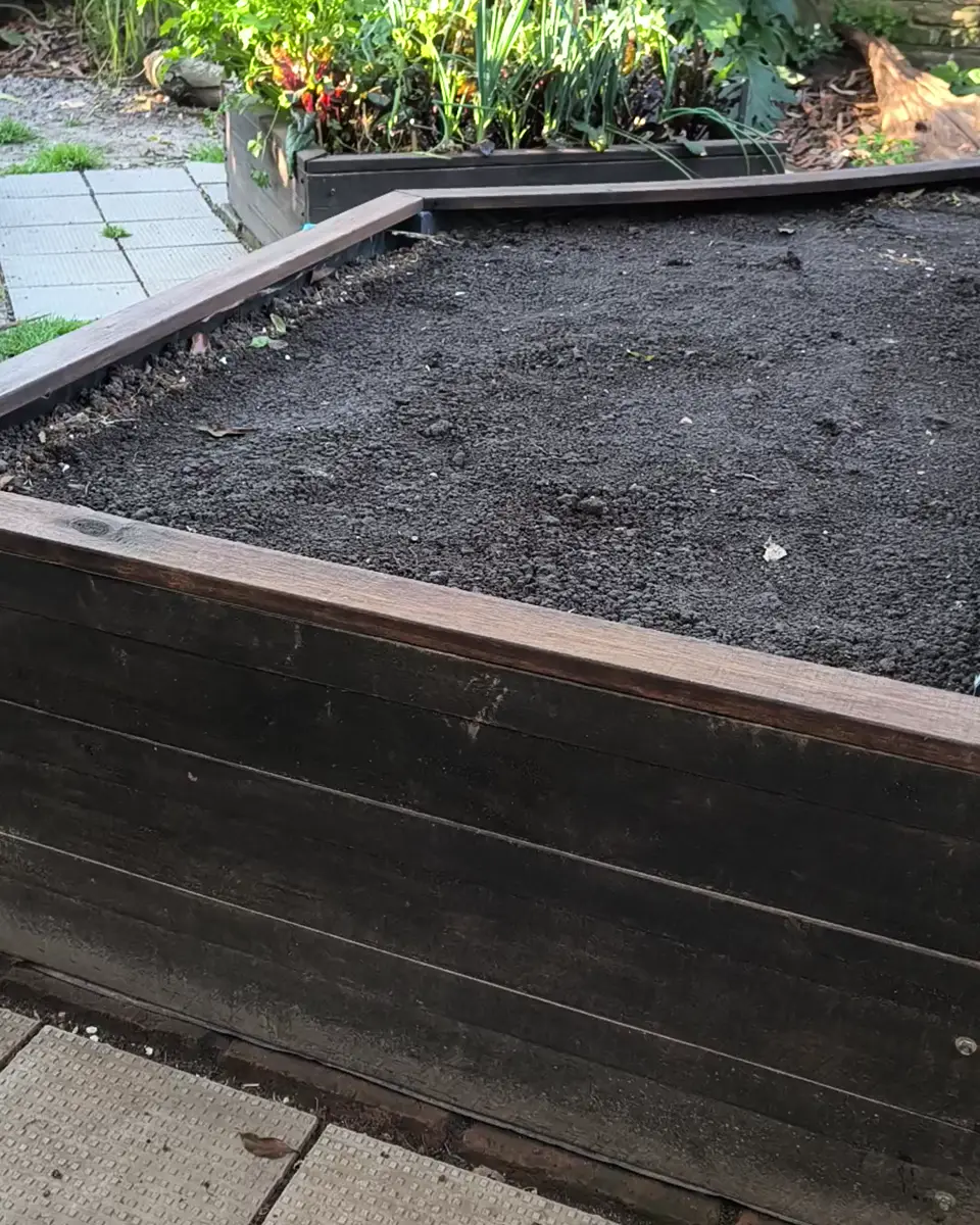 El bancal elevado de madera terminado, lleno de tierra y listo para sembrar, sobre un piso de baldosas, con el primer cajón y el jardín de fondo