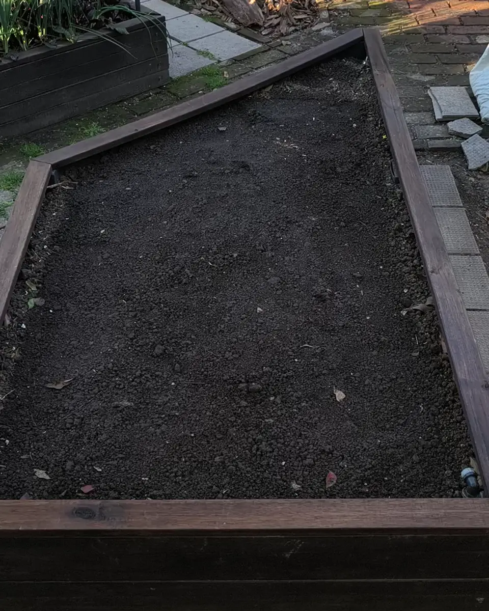 El segundo bancal elevado de madera terminado y lleno de tierra, visto desde un extremo: uno de sus lados corre en diagonal; al fondo, el primer cajón