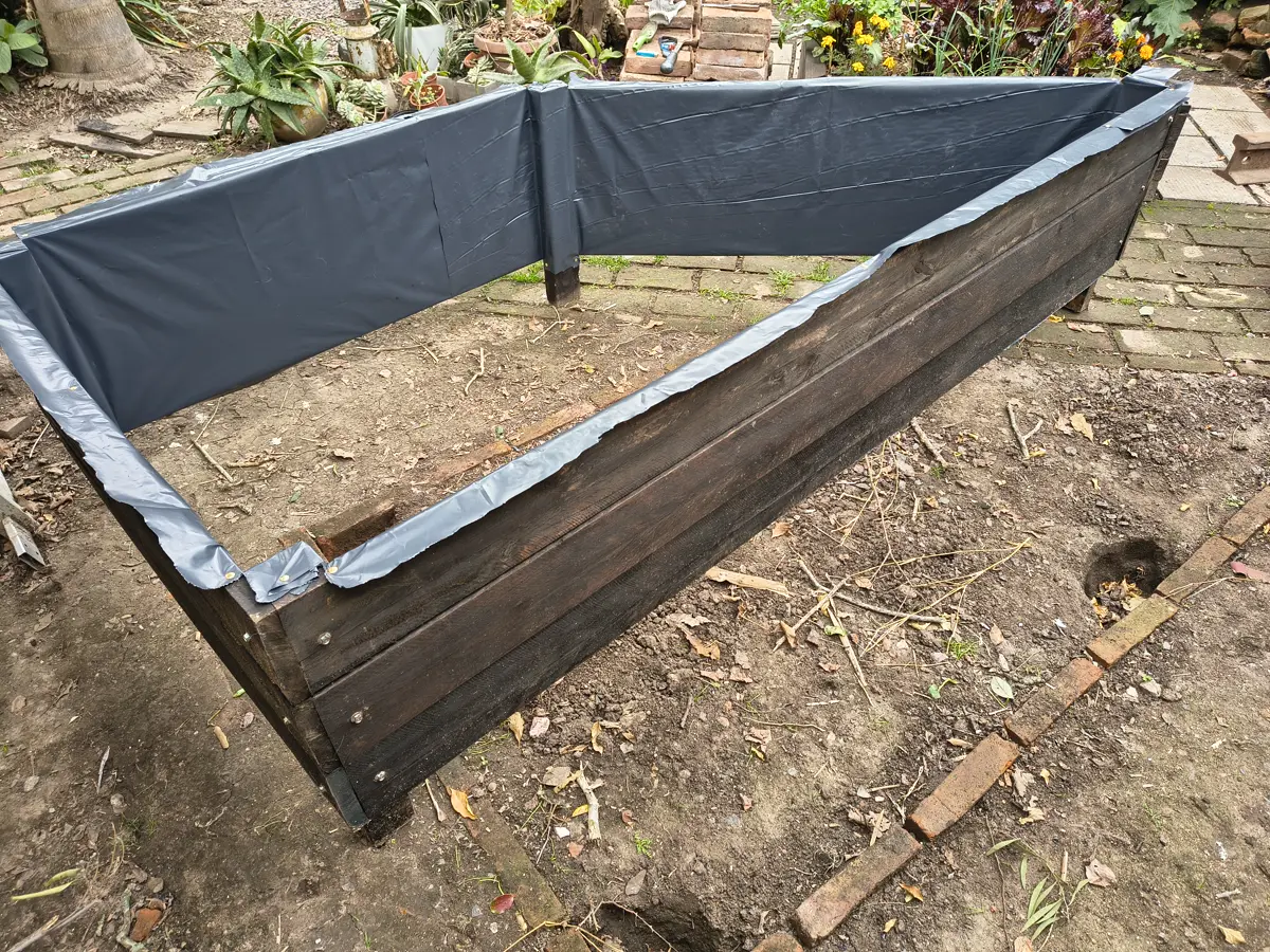 El cajón de madera oscura armado en su lugar, apoyado sobre una hilera de ladrillos, con lona plástica gris en las paredes interiores y el fondo de tierra